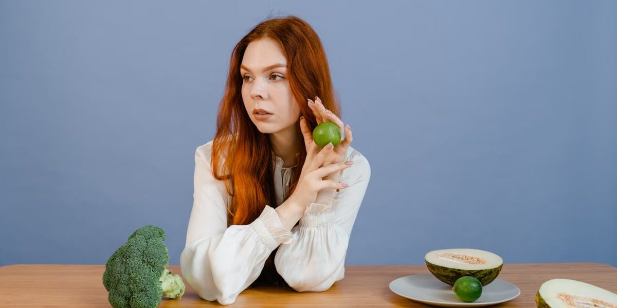 Caucasian woman sitting at table holding lime with broccoli and melon.